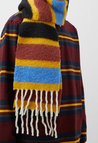 Wollschal mit horizontalen Streifen in Gelb, Blau, Schwarz und Burgund, mit Fransen an den Enden, getragen über einem gestreiften Hemd.