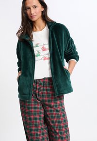 Ciemnozielona polarowa kurtka zapinana na zamek błyskawiczny z bocznymi kieszeniami, w połączeniu z białą koszulą i spodniami od piżamy w kratę w odcieniach zieleni i czerwieni.