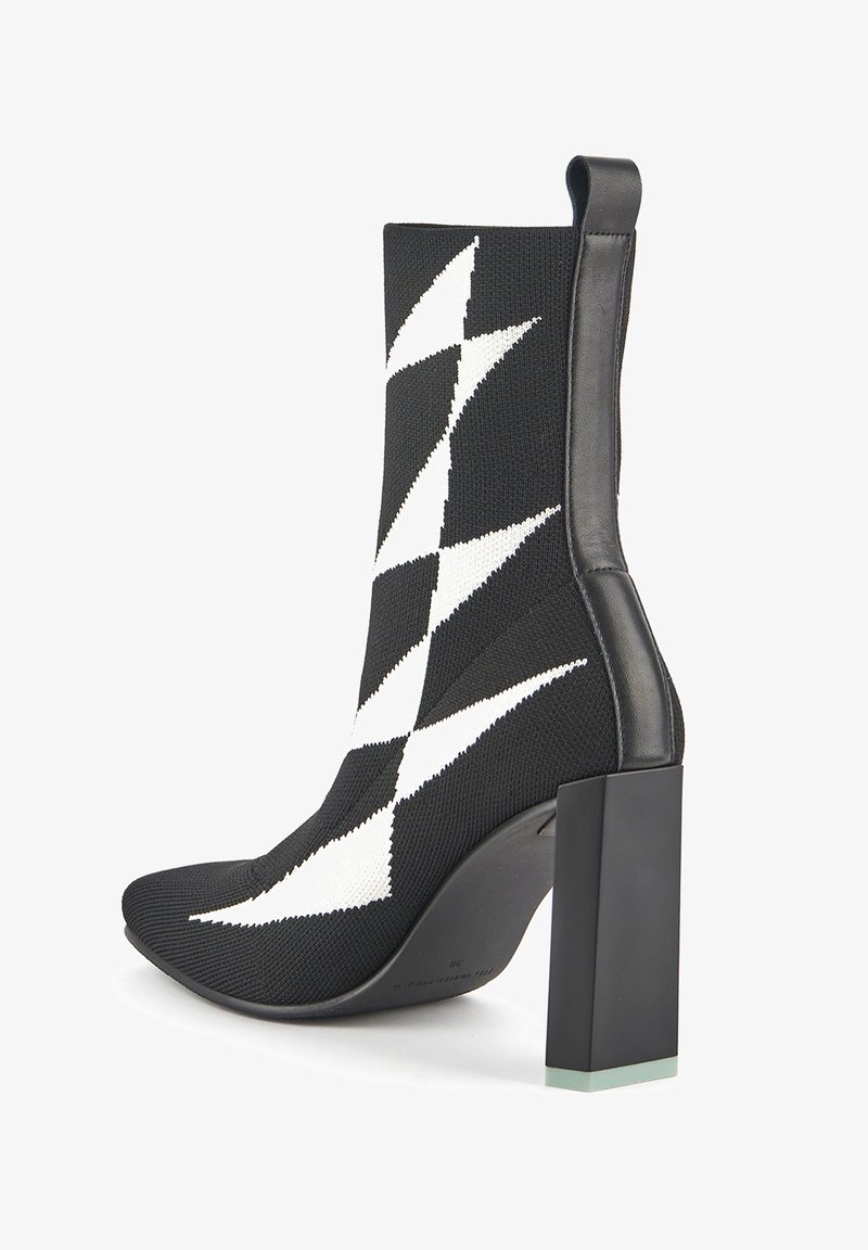 Knit ankle boot in black with white zigzag pattern, pointed toe, and block heel featuring a transparent accent at the base.
