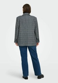 Personne portant un blazer à carreaux gris, un jean bleu et des chaussures noires, se tenant de dos contre un fond uni clair.