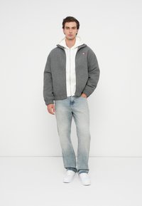 Graue Wollbomberjacke mit Reißverschluss, darunter ein weißes Kapuzensweatshirt, hellblaue Jeans und weiße Sneaker. Einfaches Design, lässige Passform.