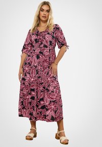 Frau mit langen blonden Haaren, die ein pinkes und schwarzes Blumenkleid in Midi-Länge und beige Plateausandalen trägt, steht vor einem schlichten hellen Hintergrund.