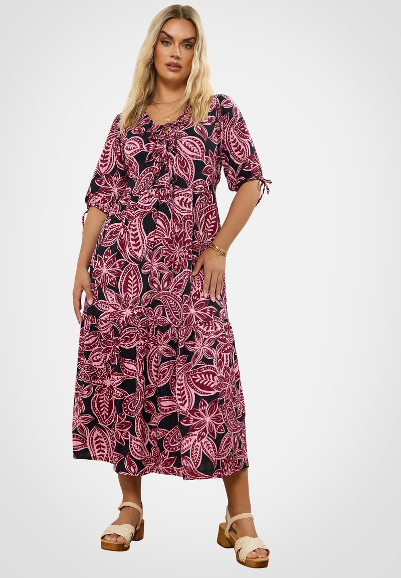 Frau mit langen blonden Haaren, die ein pinkes und schwarzes Blumenkleid in Midi-Länge und beige Plateausandalen trägt, steht vor einem schlichten hellen Hintergrund.
