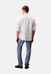 Camisa blanca de manga larga con puños enrollados, vista trasera, combinada con jeans decoloridos azules y zapatos negros. Diseño simple con un ajuste relajado.