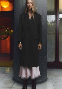 Palton lung, negru, din lână, cu umeri structurați și două buzunare frontale, purtat peste o rochie satinat roz cu dantelă. 