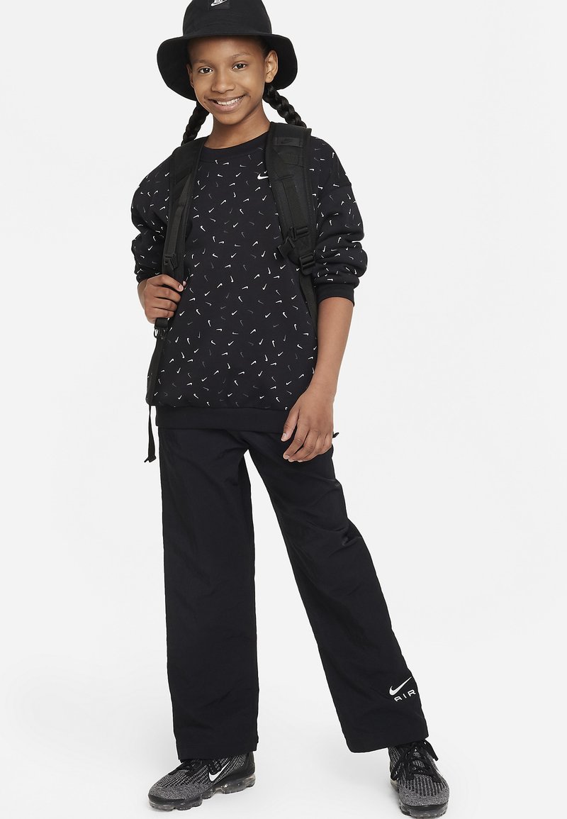 Felpa nera con motivo astratto bianco, pantaloni neri e un cappello a secchiello nero. L'outfit include uno zaino e scarpe da ginnastica nere.