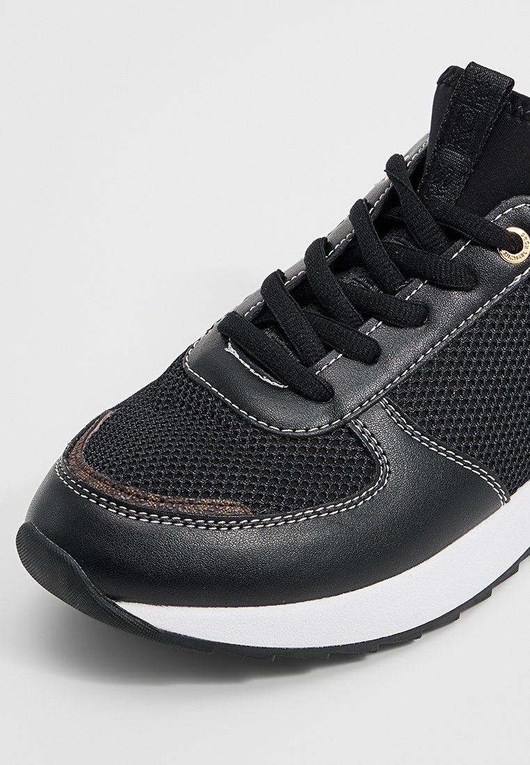 Acercamiento de una zapatilla negra con paneles de malla y cuero, cordones negros, costuras blancas y una suela blanca con suela negra.