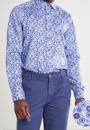 Mies, joka pukeutunut siniseen, kukkakuviolliseen pitkähihaseen paitaan, joka on sullottu tummansinisiin housuihin, pitäen toisessa kädessään vastaavan kangasnäytteen.