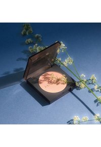 Poudre compacte dans un boîtier noir avec un couvercle transparent, contenant une poudre rose clair à l'intérieur et un logo en relief, positionnée sur un fond bleu.