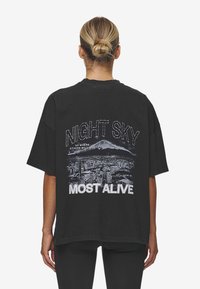Svart bomulls oversized t-shirt med ett grafiskt tryck av en stadssilhuett med berg. Texten säger "NIGHT SKY MOST ALIVE" i vitt.