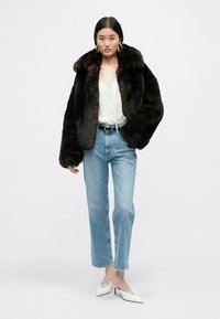 Καφέ ψεύτικη γούνα με φαρδύ γιακά, λευκό τοπ, ανοιχτό μπλε ίσια τζιν παντελόνια και λευκές μύτες με διακοσμητική λεπτομέρεια.