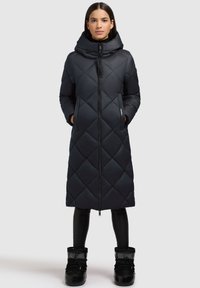 Lange, donkere, gewatteerde pufferjas met hoge kraag, rits aan de voorkant en zijzakken. Gemaakt in combinatie met zwarte, met bont gevoerde winterlaarzen.