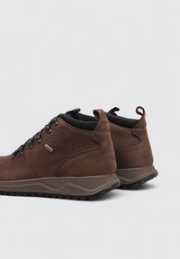 Zapatos de media caña de cuero marrón con acabado texturizado, cordones negros y suelas de goma grises. Presentan perforaciones y una pestaña para tirar en la parte trasera.