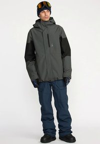 Kurtka narciarska w ciemnoszarym i czarnym kolorze, wyposażona w kieszeń na zamek, regulowany kaptur i mankiety przeciwwiatrowe. W zestawie z niebieskimi spodniami narciarskimi i czarnymi butami.