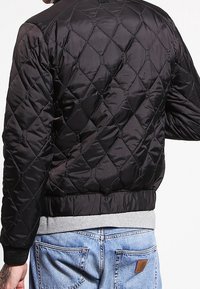 Svart vadderad bomberjacka med metallisk lyster, med ribbad krage och muddar samt en grå elastisk midja.