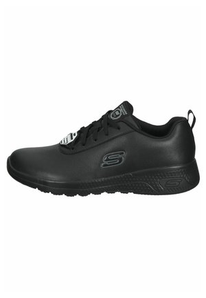 Chaussure de sport noire avec un matériau synthétique lisse, un embout arrondi, un col rembourré et une semelle extérieure texturée, avec un logo sur le côté.