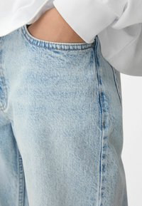 Ruka je zakopčana u prednjem džepu svijetloplavih traperica, koje su uparene s bijelom bluzom dugih rukava.