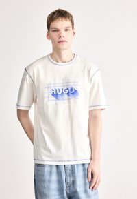 T-shirt creme com costura azul, apresentando um design gráfico azul "HUGO" num padrão de grade. Decote redondo e mangas curtas, corte descontraído.