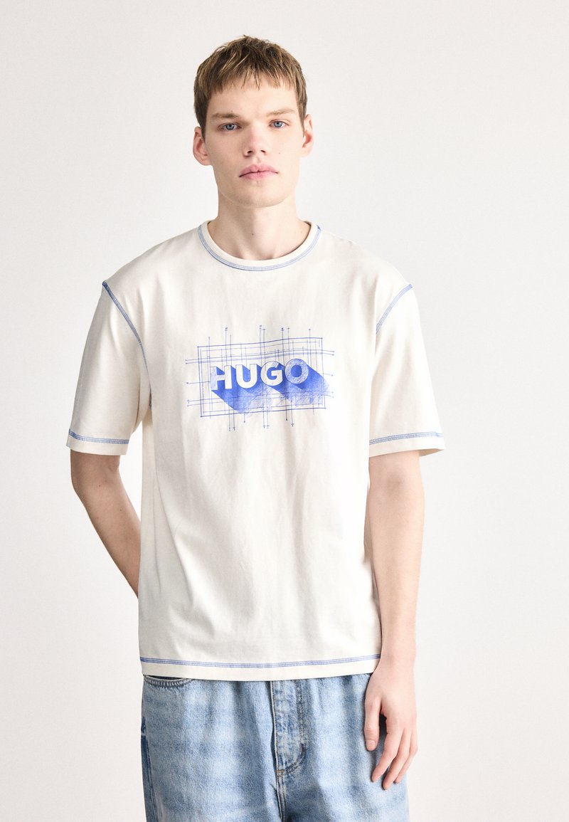 T-shirt creme com costura azul, apresentando um design gráfico azul "HUGO" num padrão de grade. Decote redondo e mangas curtas, corte descontraído.