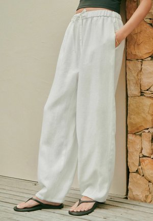 Person trägt lockere weiße Leinenhose mit elastischem Bund und schwarzen Flip-Flops, steht vor einer beigen Wand und einem Steinsäule.