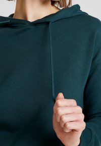 Sudadera de un azul oscuro con textura, hecha de un tejido suave, con un bolsillo frontal y capucha ajustable con cordones. Mano sujetando el dobladillo.