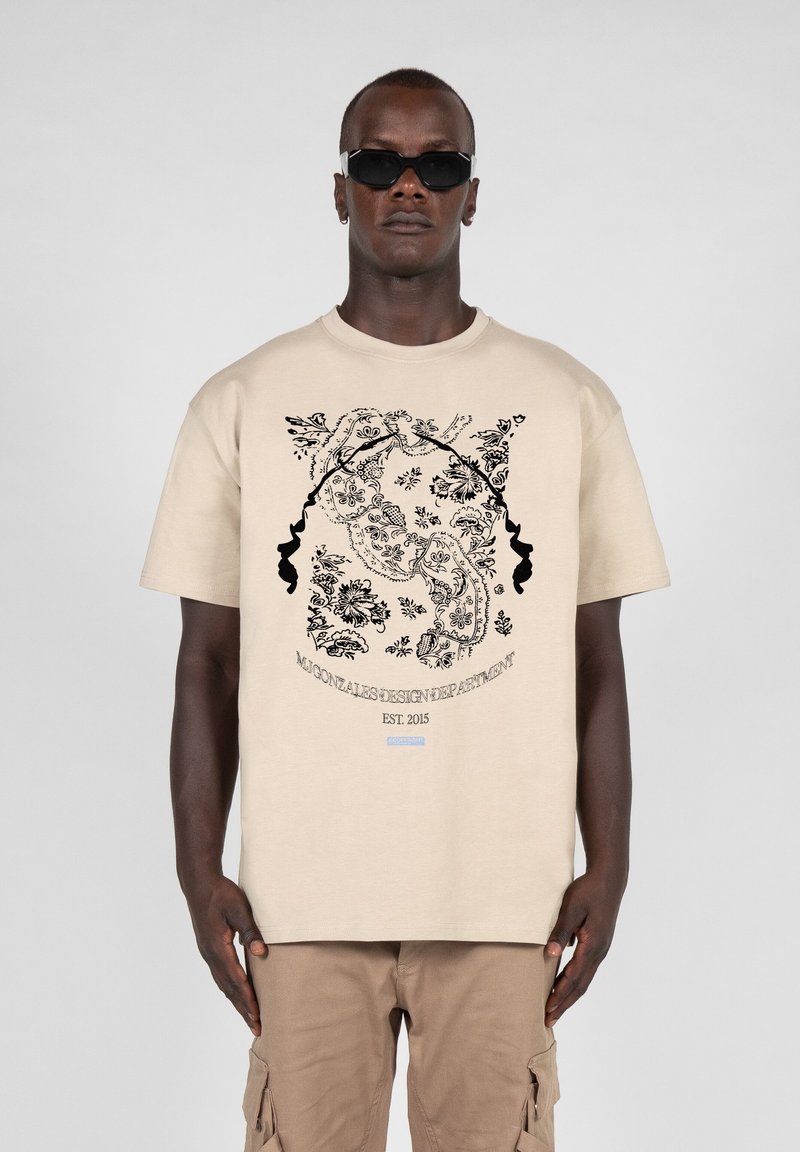 Beige bomulls t-skjorte med svart blomsterdesign og logo "Gonzalez Design Department EST. 2015". Modell som har på seg solbriller og beige cargo shorts.