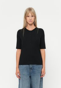 Svart kortermet topp med rund hals, med en rad knapper foran, laget av teksturert stoff. Kombinert med lyseblå vide jeans.