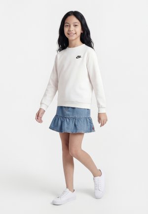 Fille aux cheveux noirs portant un sweat-shirt blanc Nike, une jupe en denim bleue avec des volants, et des baskets blanches, marchant avec un sourire sur fond blanc.