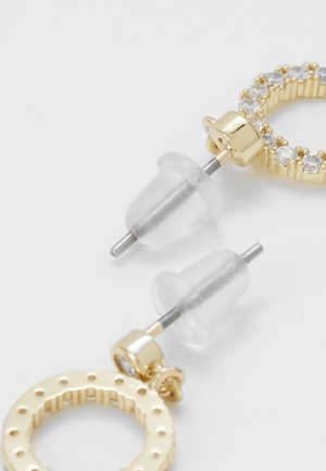 Kultaiset pyöreät korvakorut, joissa toisessa on kirkkaat kivet reunoilla ja molemmissa kiinnikkeissä läpinäkyvät silikonitaustat.