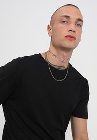Svart t-skjorte med korte ermer, tettsittende snitt. Gullkjede, synlige tatoveringsdesign på nakken med piggtråd og dekorative elementer.