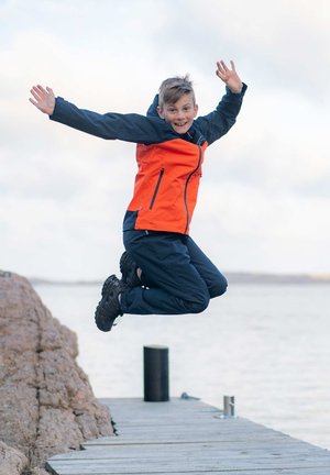Skinnende orange og marineblå jakke med lynlås og lommer, parret med mørke bukser og sorte vandtætte støvler, iført af en person, der hopper på en kaj.