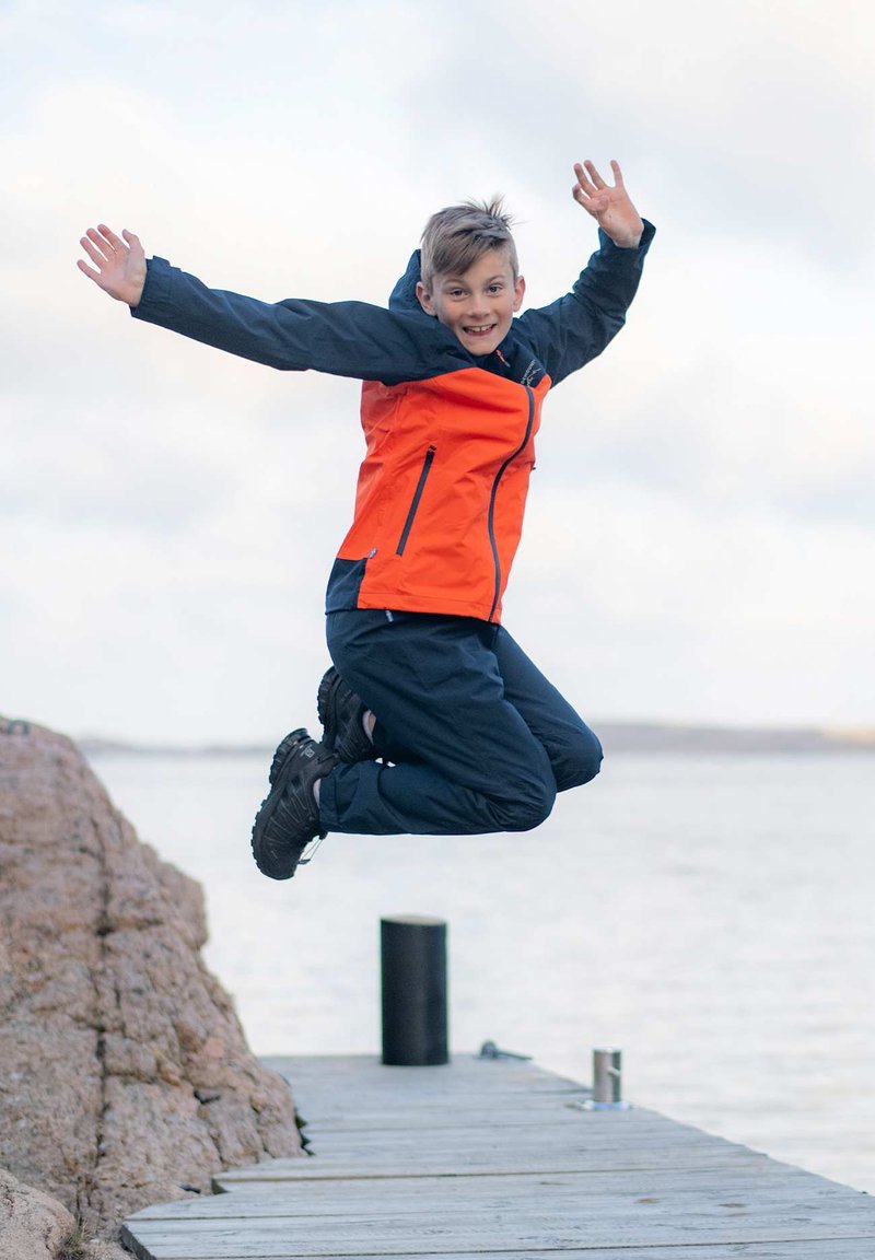 Ljus orange och marinblå jacka med blixtlås och fickor, kombinerad med mörka byxor och svarta vattentäta kängor, bärs av en person som hoppar på en brygga.