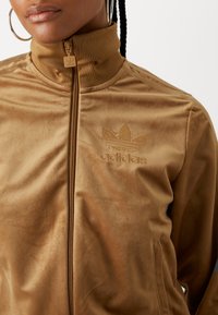 Braune Zip-Jacke aus glattem Material. Merkmale einen hohen Kragen, ein gesticktes Adidas-Logo und das klassische Drei-Streifen-Design auf den Ärmeln.