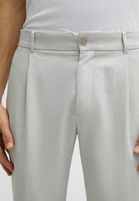 Pantaloni grigi chiari realizzati in un tessuto texturizzato, con chiusura frontale a bottone e pinces laterali. Il modello sta tenendo in mano il giro vita.