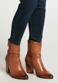 Femeie îmbrăcată în blugi skinny bleumarin închis �și cizme cu toc mediu din piele maro, care se termină la jumătatea gambei, cu fermoare laterale și curele.