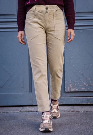 Une personne marche en pantalon en velours côtelé beige à grandes poches et en baskets épaisses, portant un pull bordeaux, devant une porte bleue.
