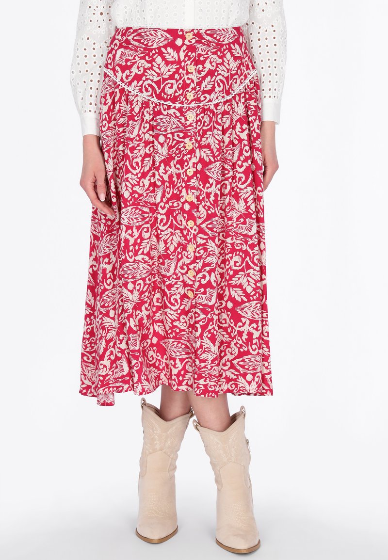 Roter Midirock mit weißem Blumenmuster und vorderen Knöpfen, kombiniert mit beigen Cowboy-Stiefeln und einer weißen Bluse mit Lochstickereiärmeln.