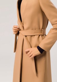 Gabardina beige con un acabado texturizado, cinturón en la cintura y mangas largas, que presenta un patrón sutil y un detalle en azul marino en los puños.