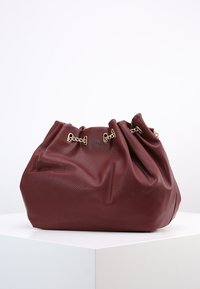 Miękka bordowa skórzana torba z zebranym wykończeniem u góry oraz złotymi akcentami łańcuszka, spoczywająca na białym piedestale na jednolitym tle.