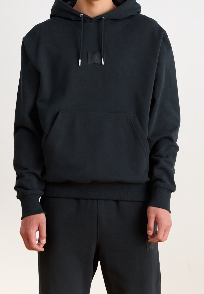 Homme portant un sweat à capuche noir et un pantalon noir assorti, debout les mains détendues le long du corps devant un fond uni.