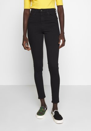 Vaqueros ajustados de cintura alta en negro, con cinco bolsillos y cierre de botón. Combinados con zapatos tipo slip-on multicolores.