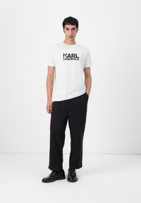 Hvit bomulls T-skjorte med svart "KARL LAGERFELD" grafikk, kombinert med svarte vide bukser og svarte sko, modell som står foran hvit bakgrunn.