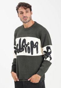 Maglione lavorato a maglia verde scuro con una striscia orizzontale bianca e testo e dettagli in stile graffiti neri sulle maniche. Polsini e orlo a coste.