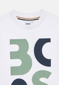 Λευκό βαμβακερό μπλουζάκι με μεγάλους πράσινους και ναυτικού μπλε αριθμούς "3" και "0" σε υφασμένο τύπωμα. Η ετικέτα BOSS είναι ορατή στο λαιμό.