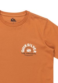 Krátký rukáv oranžového trička z bavlny se bílým kruhovým logem a textem "Quiksilver" a "The Original Boardriding" níže.
