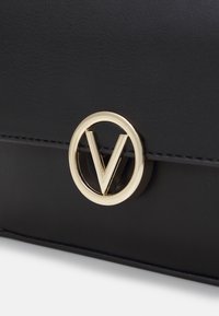 Schwarze Kunstledertasche mit einem auffälligen goldenen runden Logo mit einem 'V'-Ausschnitt. Glatte Textur und sichtbare Nähdetails.