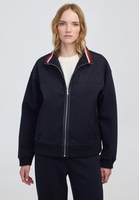 Chaqueta de navy con cremallera y cuello a rayas rojas y blancas. Tejido suave, corte relajado, logo bordado en el pecho, puños acanalados y dobladillo.