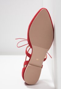 Chaussure plate en daim rouge à bout pointu avec des brides à la cheville, vue par le dessous avec la taille 37 marquée sur la semelle sur un fond blanc.