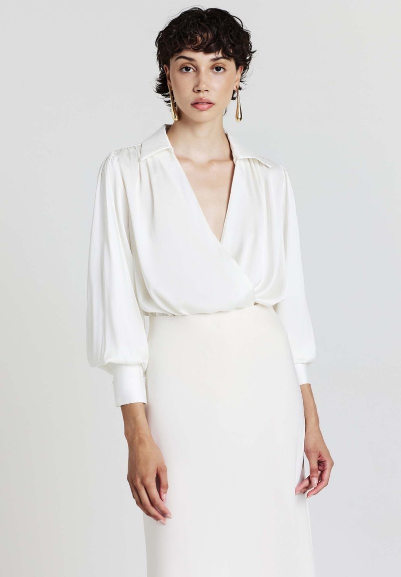 Blusa bianca a maniche lunghe con scollo a V profondo e un taglio morbido, caratterizzata da una texture setosa e delicate pieghe sulle spalle.