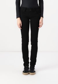 Person in schwarzen Skinny Jeans, schwarzem Langarmshirt und schwarzen Sneakers mit weißen Streifen, der vor einem schlichten weißen Hintergrund steht.